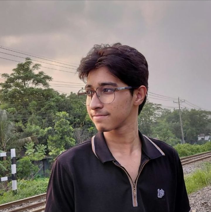Portrait of Shazzad Sayem wearing glasses and a black polo, standing by a railway track at dusk